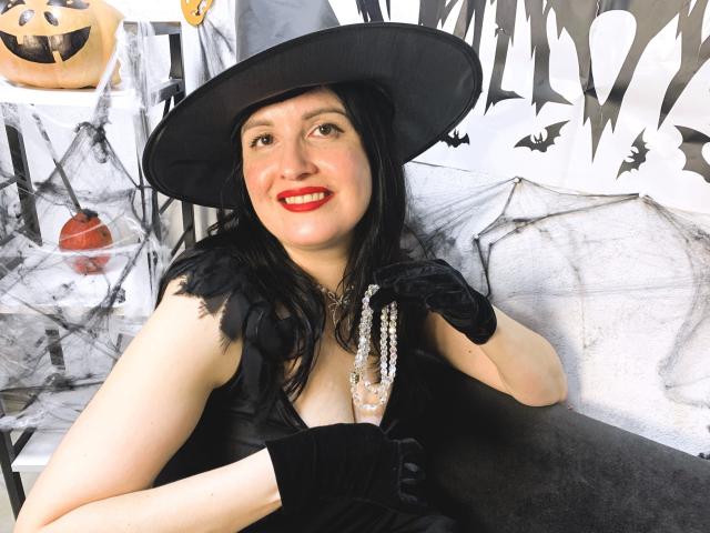 TayaBoret with black hat, red lipstick, black dress, and gloves, holding beaded necklace, smiling, showing tits, sitting, black hair, white background, Halloween theme.