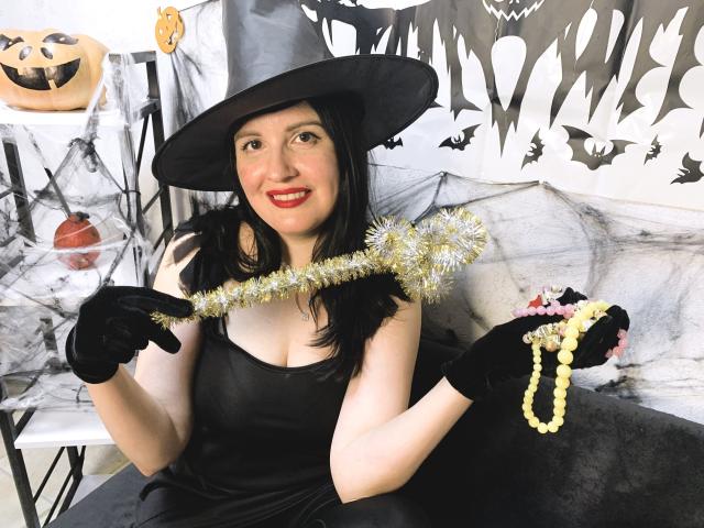 TayaBoret with black dress, hat, and gloves, holding tinsel and beads, smiling, showing red lipstick, pale skin, and black hair, with visible tits, in Halloween costume.