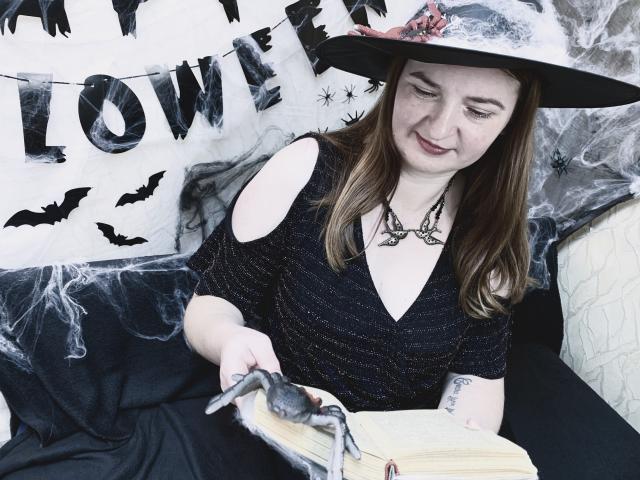 FreyaDak with brown hair, black dress, and hat, holding a gray spider on an open book, smiling. She's got a tattoo on her right arm and a necklace.