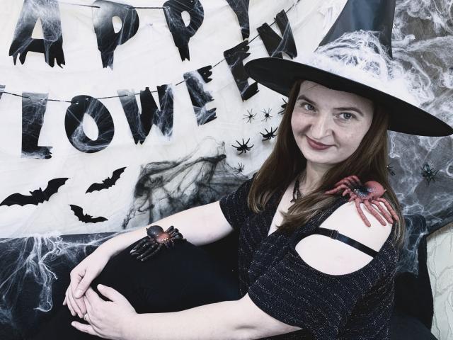 FreyaDak with pale skin, brown hair, and a witch hat, wearing a black dress with cutouts, showing a red spider tattoo on her right arm, smiling.