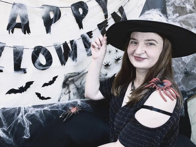 FreyaDak with long brown hair, wearing a black witch hat and cut-out black top, has a spider tattoo on her right shoulder, smiling at the camera.