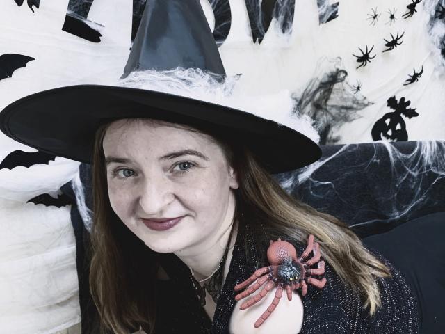 FreyaDak with a witch hat, smiling, has a spider brooch on her black, sparkly shirt, and pale skin, with brown hair, wearing dark lipstick.