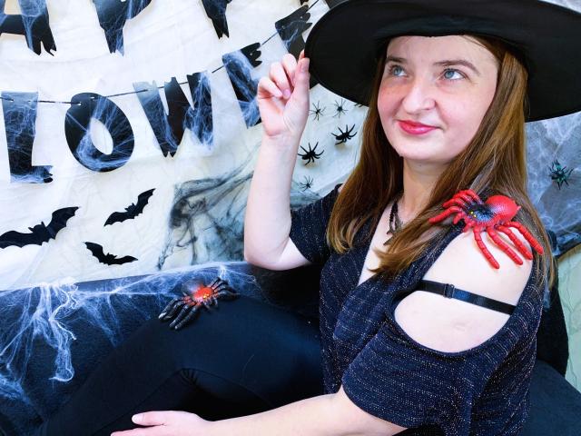 FreyaDak, com uma tatuagem de aranha vermelha no ombro, um chapéu preto e uma blusa com um ombro à mostra, está sentada sorrindo, exibindo a mão, vestindo um traje com tema de Halloween, com calças pretas e decoração de aranha..