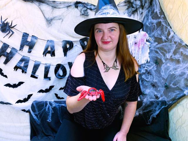 FreyaDak with red painted nails, black off-shoulder top, witch hat, necklace, holding red string, smiling, medium tits, fair skin, brown hair, Halloween setup.