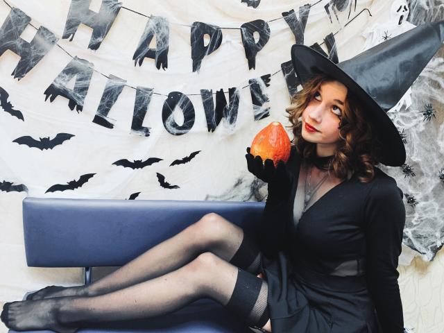 LettyCurly with curly brown hair, wearing a black witch hat, dress, and stockings, holding a pumpkin, sitting on a blue bench, black gloves, looking to the side.