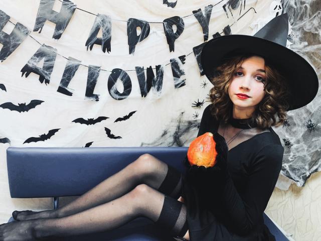 Letty Curly, gekleed als een heks, zittend op een blauwe bank met haar benen gekruist, houdt een pompoen vast, draagt zwarte kousen en een zwarte hoed, in een Halloween-thema decor..