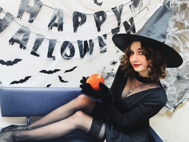 LettyCurly with curly brown hair, wearing a black witch hat, dress, and gloves, holding an orange pumpkin, sitting on a blue bench, showing her tits and ass.