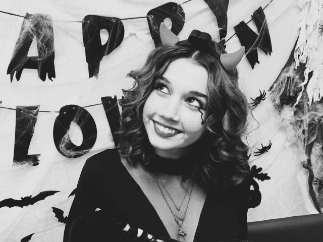 Black-and-white photo of LettyCurly with curly hair, wearing a black top, devil horns, and a choker, smiling at the camera, showing her teeth, with bats in the background.