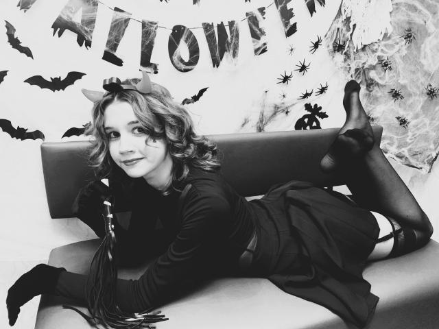 LettyCurly with curly hair, wearing a dark dress, black stockings, and bat ears, lying on a bed, legs up, showing her bare ass, and a whip beside her.