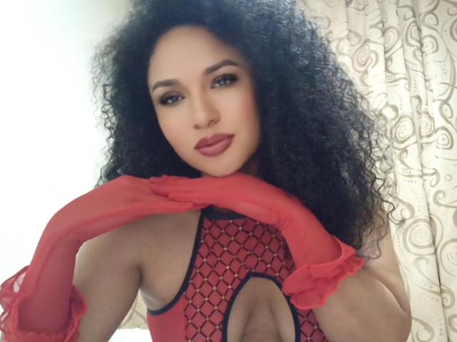 CarlaMuille with curly black hair, wearing red lace-up bodysuit and matching red gloves, holding her neck, looking at camera with a slight smile, showcasing her tits.