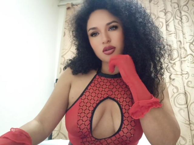 CarlaMuille with curly black hair, wearing a red, cut-out, patterned dress, exposing her tits, and red gloves, touching her chin, with a seductive look, in a photo.