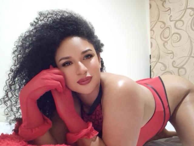 CarlaMuille, con i capelli neri ricci, rossetto rosso e lingerie rossa, indossa guanti rossi, è sdraiata sul letto, guarda la fotocamera, ha le mani sul viso, mostrando il suo seno lucido, abbronzato e formoso..