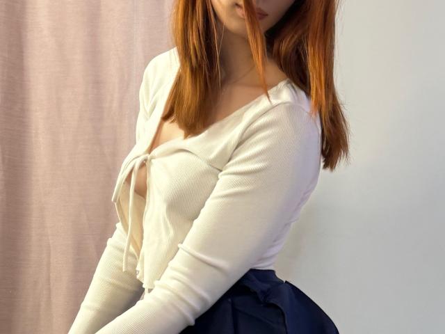ElfinVibes with red hair, white knotted top, showing small boobs, and blue skirt, standing with head tilted, mouth slightly open.