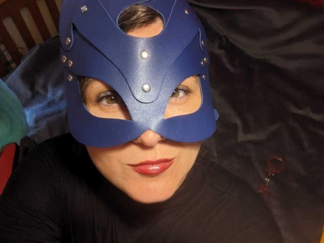 PlayRealMe trägt eine blaue Maske mit Metallnieten, roten Lippenstift und ein schwarzes Hemd, liegt auf dunklem Stoff, blickt mit grünen Augen in die Kamera, wobei die Brüste leicht sichtbar sind..