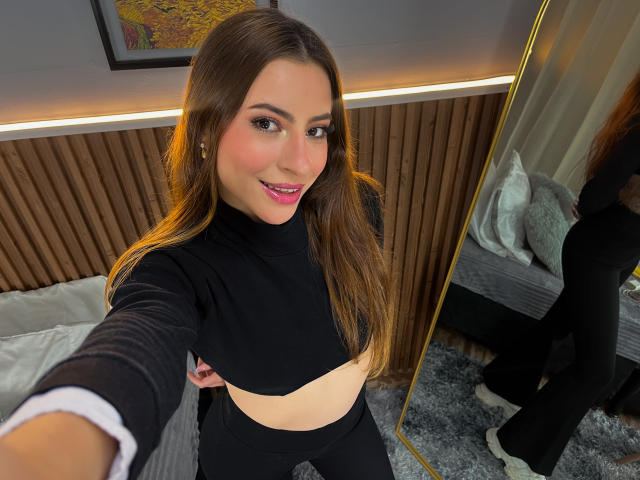 DaphneBlakee with long brown hair, pink lipstick, and black crop top, showing her midriff, taking a selfie in a bedroom with a mirror reflecting her back.