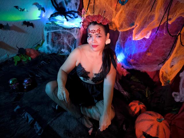 AbriilBeltran, dressed in a black lace top and skirt, sits with her tits visible, wearing a red henna forehead design, and a flower crown, surrounded by Halloween decorations.
