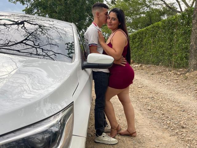 KattaAndJhon, standing by a white car, kissing. KattaAndJhon, curvy with big ass, in red dress, holding Jhon's arm. Jhon, tall, slim, white shirt, dark pants.