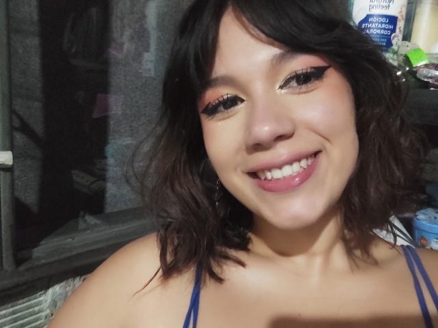 MillyJohnson has short black hair, pink eyeshadow, thick black eyeliner, and a big smile showing white teeth. She's wearing a blue strap top, and has a cute face.