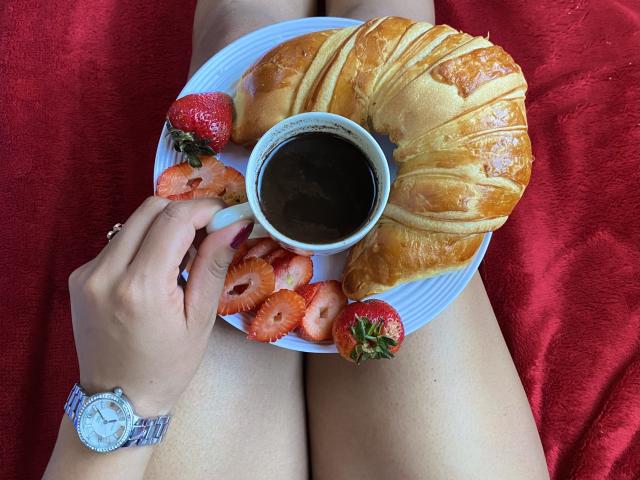 SophieSonyas's hands holding a white plate with a flaky croissant, black coffee, sliced strawberries, and a strawberry. Red blanket under her, wristwatch on her left hand.