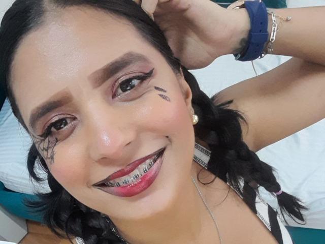 O rosto de Maria Smith, com tranças escuras, sombra rosa, delineador preto, batom vermelho, sorrindo com aparelho ortodôntico, tatuagens pretas nas bochechas, uma pulseira azul e brincos dourados..