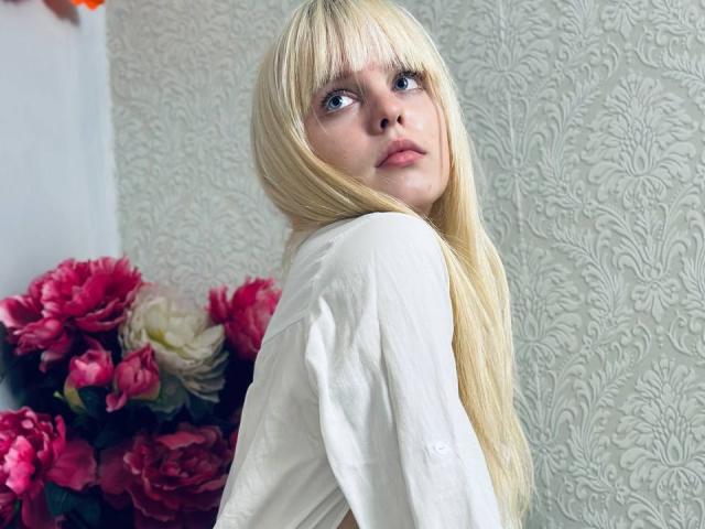Lialit mit langen blonden Haaren, blauen Augen und einem weißen Hemd, die über die Schulter schaut, hat ein süßes Gesicht und kleine Brüste. Im Hintergrund sind rosa und weiße Blumen zu sehen..