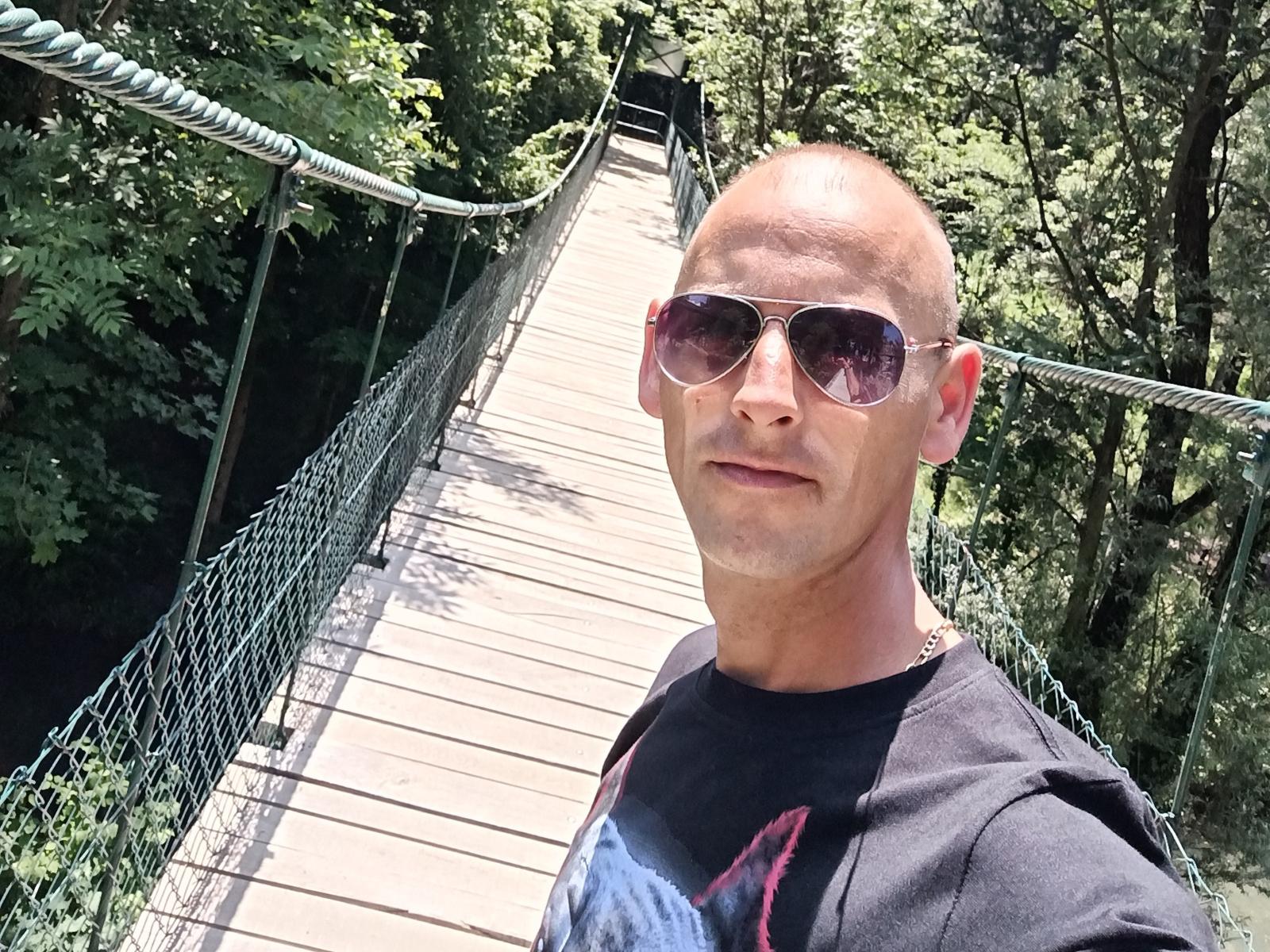 Cirkon69 with bald head, wearing pink-tinted sunglasses and black shirt, standing on a wooden suspension bridge with green netting, surrounded by trees in bright sunlight.