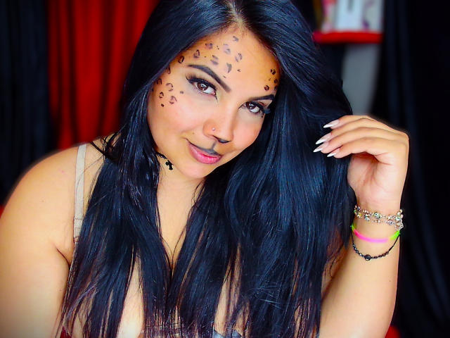 AvaChanne with long black hair, leopard print face paint, and a nose ring, smiling at the camera, showing off white nails and a bracelet. Boobs, cute, and sexy.
