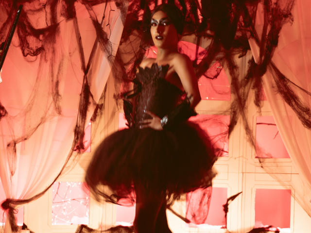 FallonRogers in a black, strapless, frilled dress with a fluffy skirt, standing in front of red and black sheer curtains, wearing dark makeup and a watch.