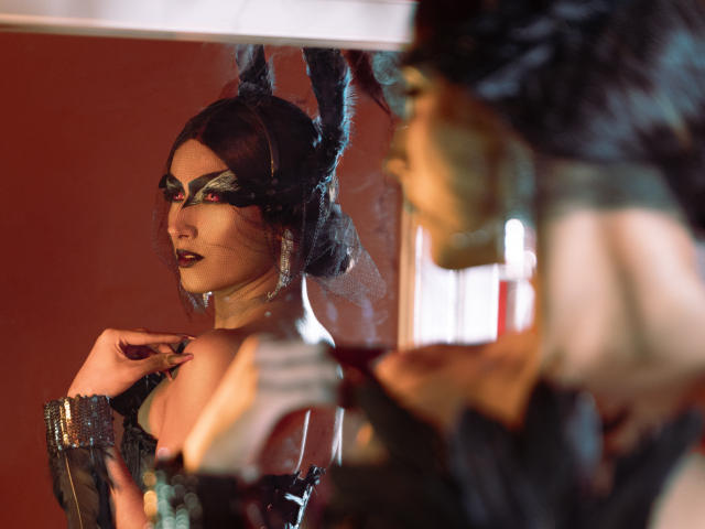 FallonRogers, con maquillaje oscuro, encaje negro y un tocado con plumas, mirando fijamente al espejo. El reflejo muestra su rostro, una mano con un guante sin dedos y su cabello oscuro, todo iluminado por una luz suave..