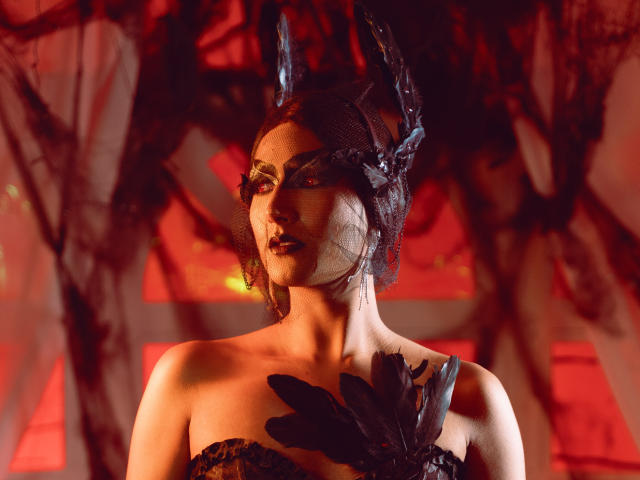 FallonRogers, avec un maquillage sombre, une pièce de tête en dentelle noire et une poitrine ornée de plumes noires, se tient sous une lumière rouge, avec une expression intense. Ses seins sont couverts de dentelle noire et de plumes..