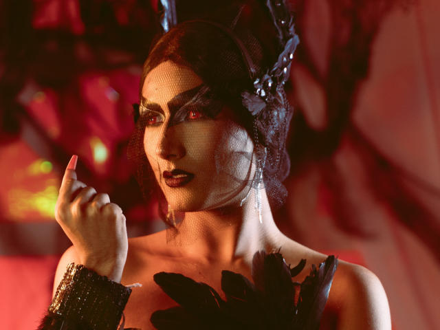 FallonRogers with dark makeup, red eyes, black feather headpiece, and wrist cuff, holding finger to mouth, showing off bare shoulders and black feathered accessory.