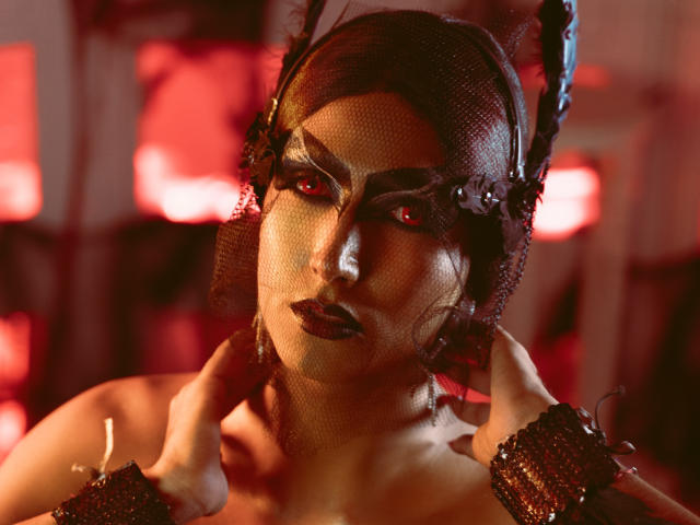 FallonRogers with dark red eyes, wearing a black mesh headpiece with feathers, textured gold makeup, and black wrist cuffs, holding her head with hands, in a red-lit scene.