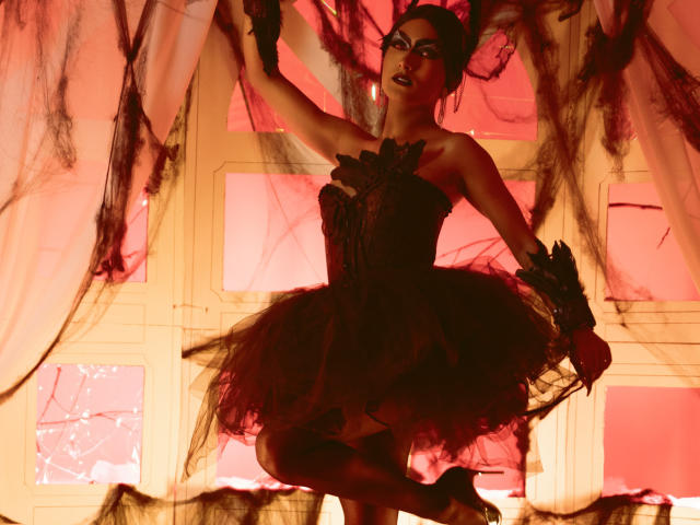 FallonRogers in a black, lace-up, strapless ballgown with a fluffy tutu, black gloves, and dark makeup, standing with arms raised, holding sheer black curtains, in a red-lit room.