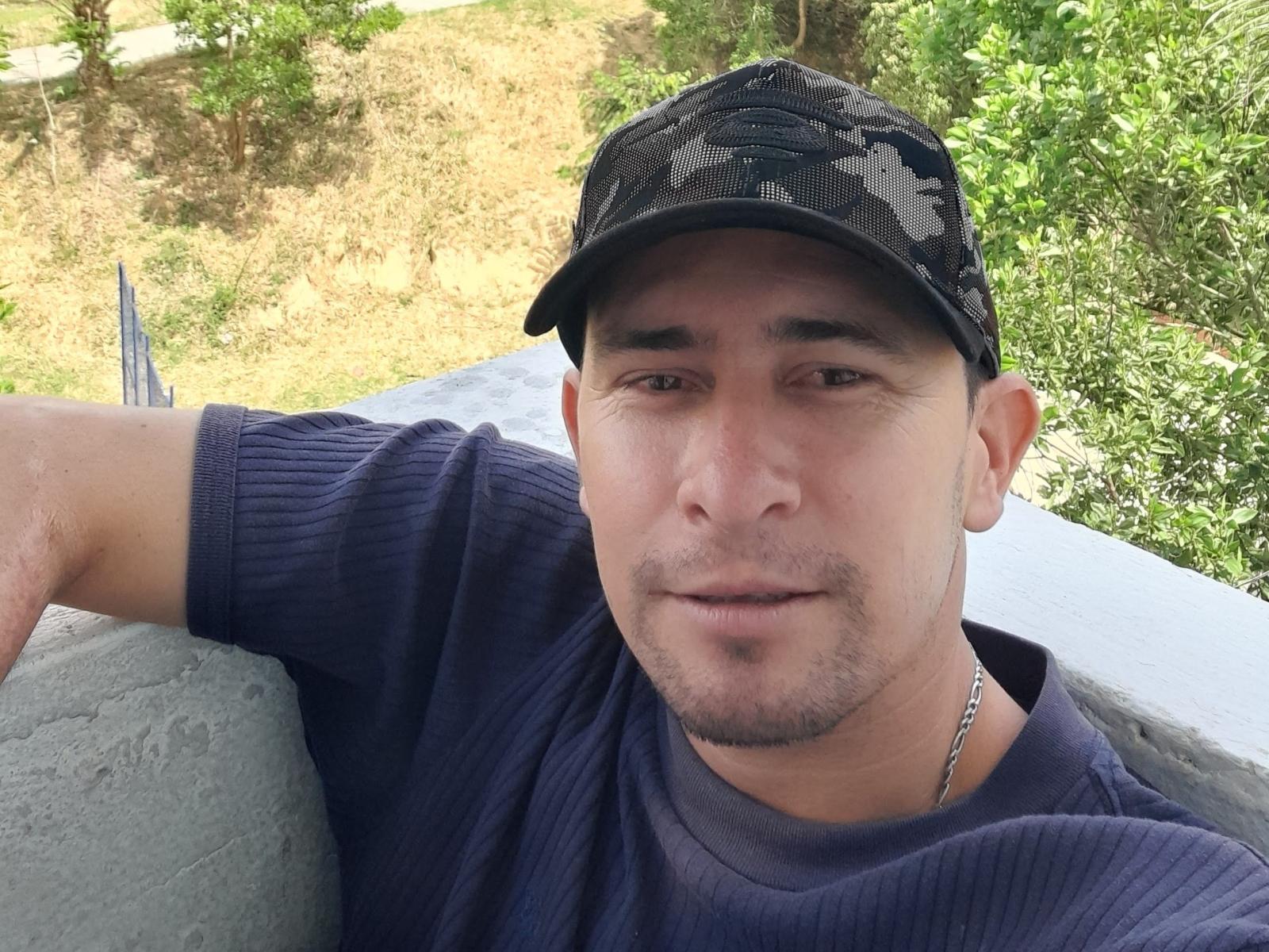 CrisHotBoy with a camo baseball cap, short dark hair, stubble, and a slight smile, wearing a purple ribbed shirt, leaning on a white wall.