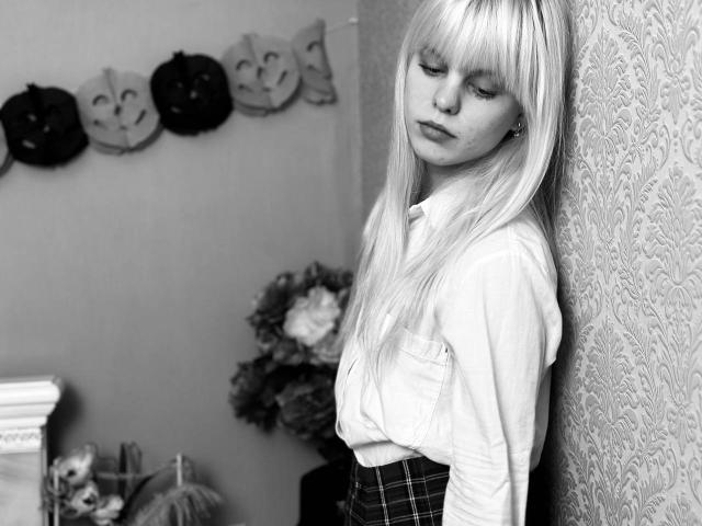 Lialit with long blonde hair, eyes closed, wearing a white shirt and plaid skirt, leaning against a patterned wall, showing a serious, slightly sad expression, with a small nose.
