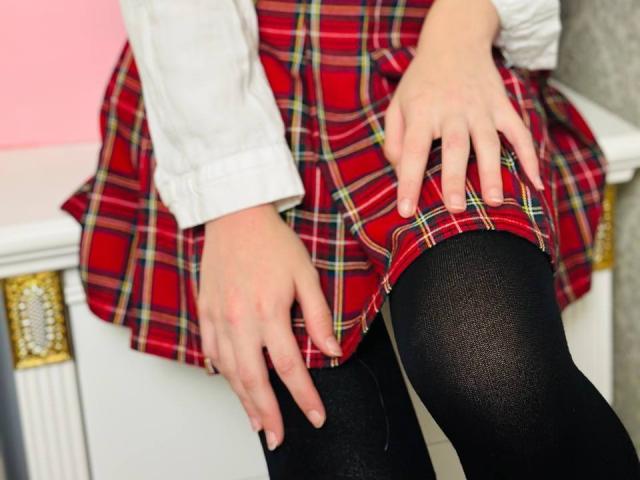 Lialits Hände ruhen auf ihrem roten karierten Rock, den schwarzen, blickdichten Strumpfhosen und dem weißen Hemd. Ihre Finger berühren ihren Oberschenkel und zeigen ein süßes, verführerisches Schulmädchen-Outfit..