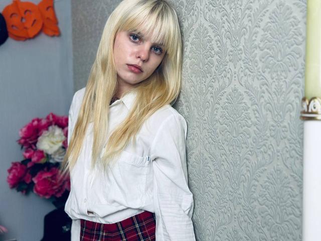 Lialit, con cabello rubio largo, ojos azules y una camisa blanca, apoyada contra una pared texturizada, vestida con una falda escocesa roja, con una expresión seria, de pie junto a flores rosas y blancas..
