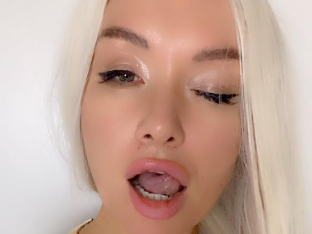 AvaGreenx has blonde hair, shiny eyes with black eyeliner, and her tongue is out, showing a small amount of saliva on her pink lips. Her face is slightly glossy.