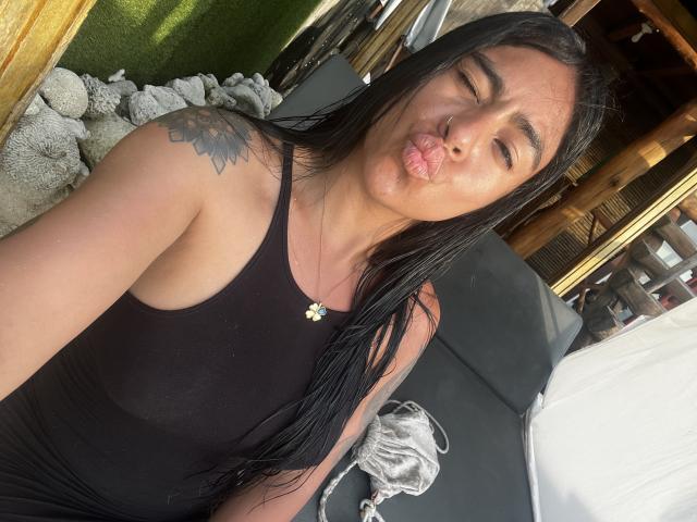 StellaBeckker with long black hair, puffy lips, black tank top, tattoo on shoulder, gold necklace, looking into camera, sitting on dark couch, white pillow in background.