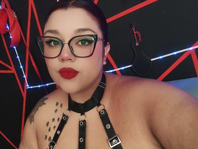 EmilyDom with glasses, red lipstick, and black harness, showing her tits, has green eye makeup, a tattoo on her shoulder, and a black ball gag on the wall.