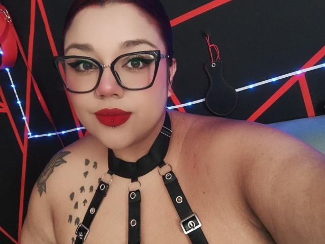 EmilyDom with red lipstick, black glasses, black harness, nipple piercings, and a tattoo on her shoulder, looking at the camera with a slight smile. Her tits are visible.