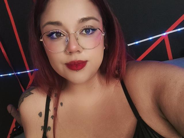 EmilyDom with red hair, glasses, nose ring, red lipstick, pierced lip, black tank top, tattoos on shoulder, looking at camera, dark background with red and blue lights.