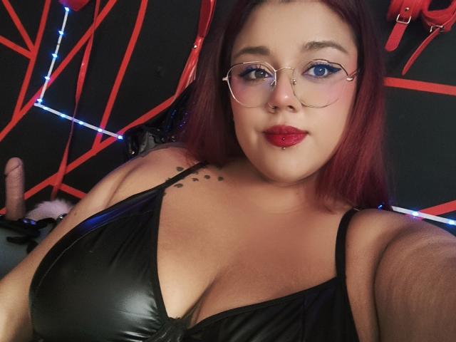 EmilyDom with red hair, glasses, and red lipstick, wearing a black leather top showing her big tits, a nose ring, and a tattoo on her right shoulder.