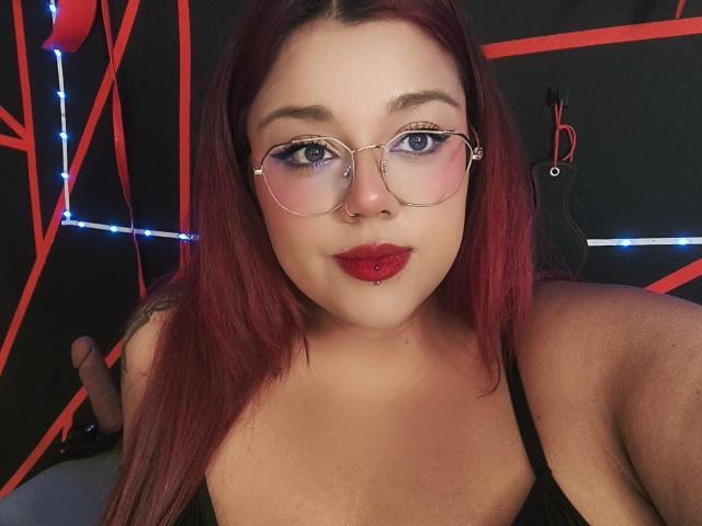 EmilyDom with red lipstick, glasses, and red hair, looking at the camera, wearing a black top, showing her tits, nose ring, and blue eyes.
