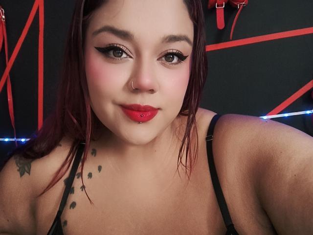 EmilyDom with red lipstick, nose ring, and lip piercing, wearing a black strap top, showing tattoos on her shoulder, and looking at the camera with black eyeliner.