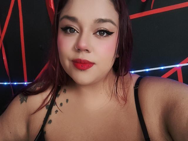 EmilyDom with red lipstick, black eyeliner, nose ring, and tattoos on her left shoulder. She's wearing a black top, her red hair down, and a subtle smile.