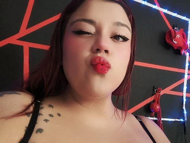 EmilyDom with red lipstick, nose piercing, and black eyeliner, pouting her pink, pierced lips, showing a tattoo on her right shoulder, wearing a black strap top.