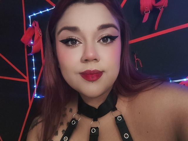 EmilyDom with red lipstick, black eyeliner, and a nose piercing, wearing a black leather choker with metal rings, and a red nose ring, has a slight smile.