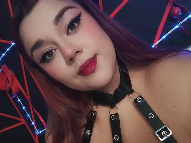 EmilyDom with red lipstick, black eyeliner, and a nose piercing, wearing a black leather harness, has pinkish-red hair, and a slight smile, showing pierced lips.