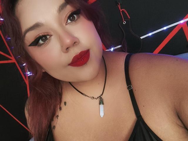 EmilyDom has red lipstick, black eyeliner, and a nose piercing. She's wearing a black top, a necklace with a white stone, and has red-dyed hair. Boobs visible.